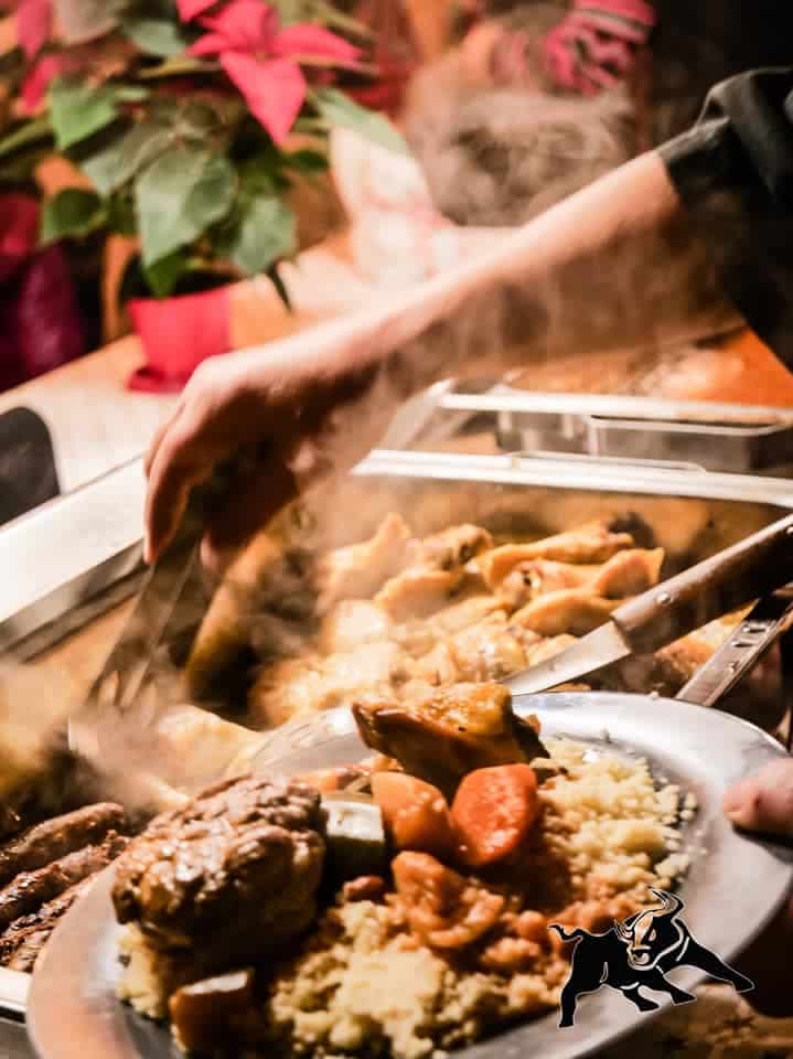 Service de buffet : couscous, viande, merguez fumantes. Une main utilise des pinces. Assiette avec logo taureau.
